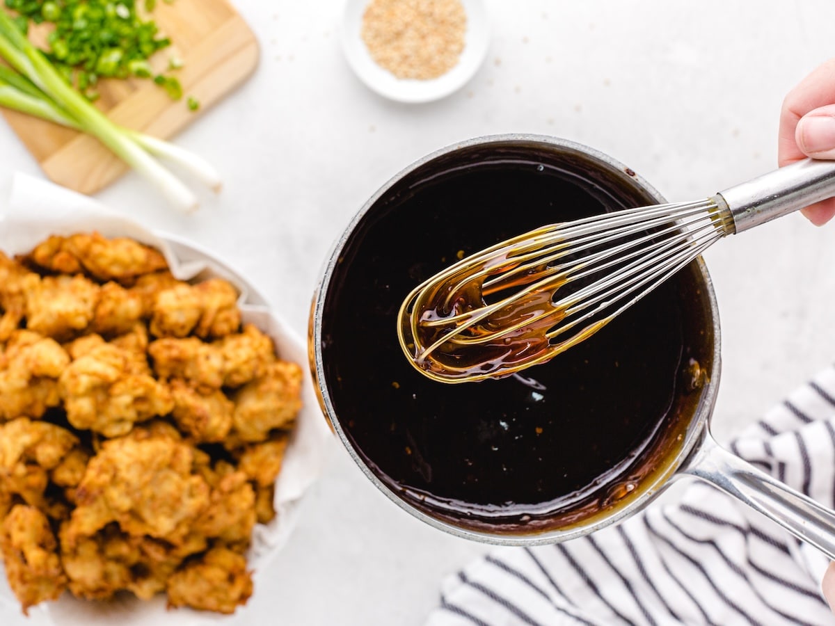 Whisking the homemade sesame sauce in a saucepan next to a bowl of crispy fried chicken pieces ready to be tossed.