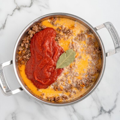 Overhead process photo of Bolognese sauce ingredients cooking together in a skillet, featuring browned ground beef, whole milk, crushed tomatoes, and a bay leaf before being stirred into the classic Italian meat sauce.