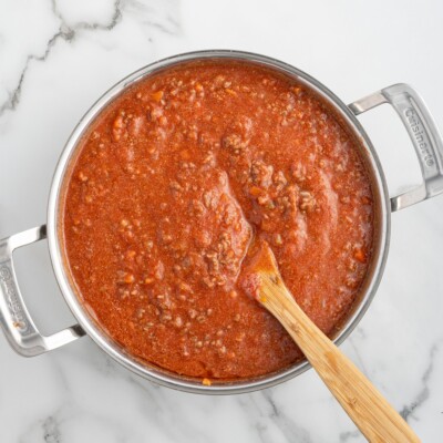 Overhead view of homemade Bolognese sauce simmering in a large skillet, showing the rich tomato-based meat sauce being stirred with a wooden spoon as the beef, tomatoes, and vegetables cook down into a thick, hearty sauce.