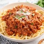 Recipe card image for homemade Bolognese featuring a bowl of spaghetti topped with rich meat sauce, grated Parmesan cheese, and fresh parsley. The photo showcases the finished Bolognese recipe served family-style with a side of Parmesan and fresh herbs.