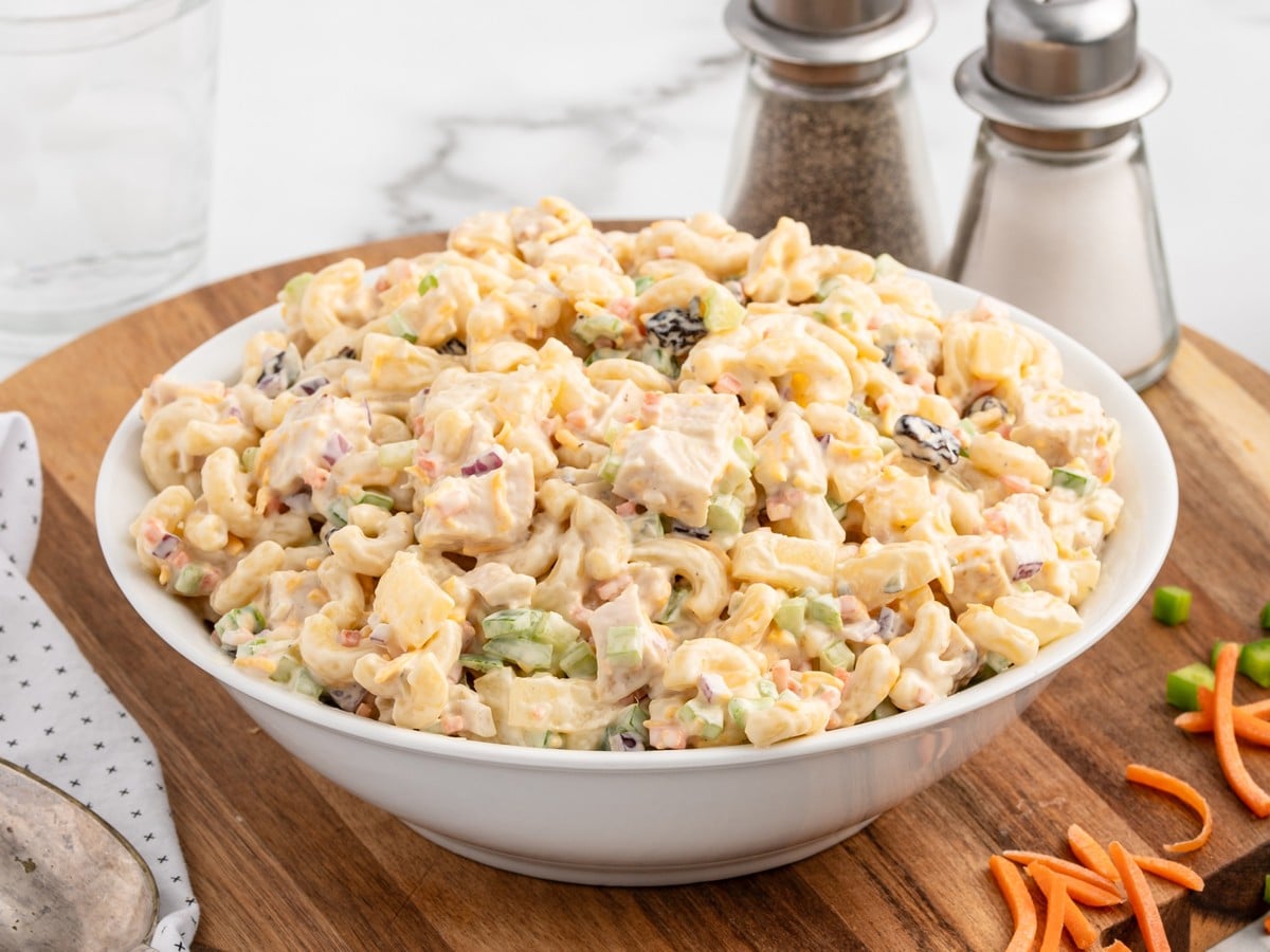 A large white bowl of creamy Chicken Macaroni Salad with elbow macaroni, chicken, pineapple, celery, carrots, red onion, raisins, and cheddar cheese on a wooden board.