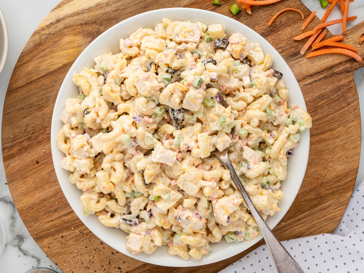 Overhead view of Chicken Macaroni Salad served in a white bowl on a wooden board, featuring tender chicken, elbow macaroni, pineapple tidbits, celery, carrots, red onion, raisins, cheddar cheese, and creamy dressing.