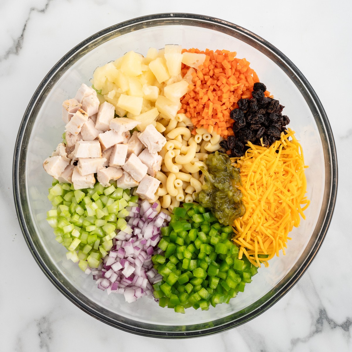 Overhead process photo showing Chicken Macaroni Salad ingredients arranged in a glass bowl, including cooked elbow macaroni, diced chicken breast, pineapple tidbits, celery, red onion, green peppers, carrots, raisins, sweet relish, and shredded cheddar cheese before mixing.