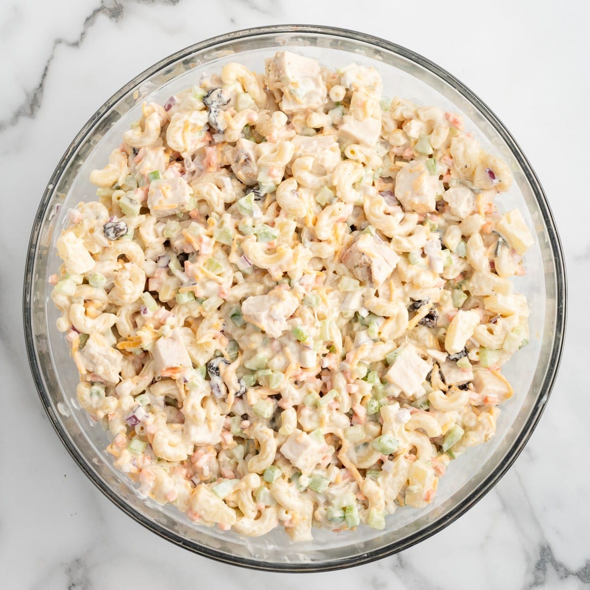 Overhead view of finished Chicken Macaroni Salad in a large glass mixing bowl, featuring elbow macaroni, tender chicken breast, pineapple tidbits, celery, carrots, red onion, green peppers, raisins, cheddar cheese, and creamy dressing.