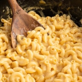 Close-up of creamy Crockpot Velveeta Mac and Cheese with a wooden spoon resting in cheesy elbow macaroni inside a black slow cooker.
