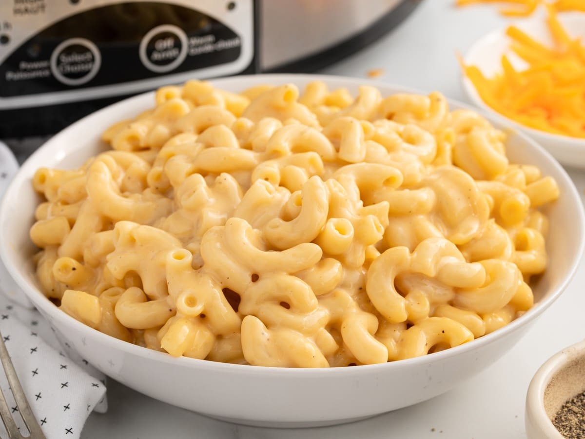 Bowl of creamy Crockpot Velveeta Mac and Cheese filled with cheesy elbow macaroni, served with the slow cooker and shredded cheddar cheese in the background.