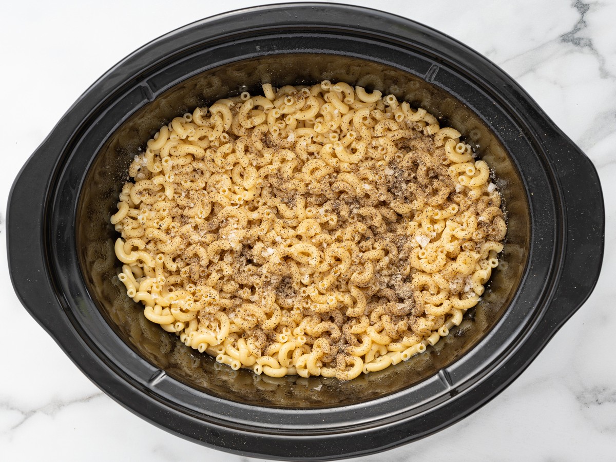Overhead photo of cooked elbow macaroni in a slow cooker seasoned with kosher salt and black pepper for Crockpot Velveeta Mac and Cheese.