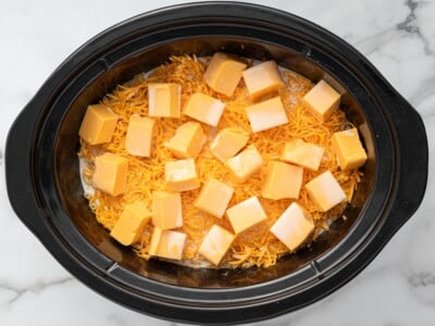 Overhead photo of cooked macaroni topped with shredded cheddar cheese, cubed Velveeta cheese, and milk in a slow cooker before cooking Crockpot Velveeta Mac and Cheese.