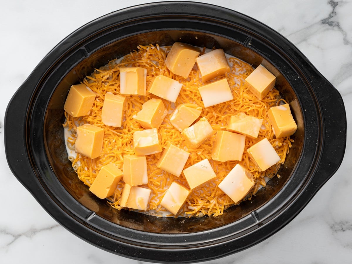 Overhead photo of cooked macaroni topped with shredded cheddar cheese, cubed Velveeta cheese, and milk in a slow cooker before cooking Crockpot Velveeta Mac and Cheese.