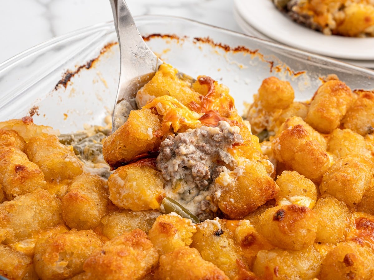 Hamburger Tater Tot Casserole being scooped from a glass baking dish with crispy tater tots, melted cheddar cheese, seasoned ground beef, green beans, and creamy filling.