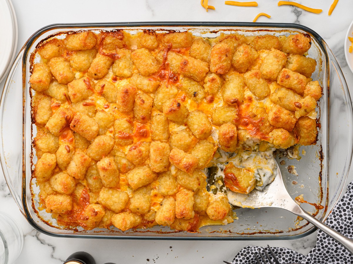Overhead view of Hamburger Tater Tot Casserole in a glass baking dish with a golden tater tot topping, melted cheddar cheese, creamy ground beef and green bean filling, and a serving spatula lifting a portion from the pan.