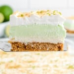 A slice of key lime lush being lifted from the pan showing the graham cracker crust, cream cheese layer, and green key lime filling topped with whipped topping and lime zest