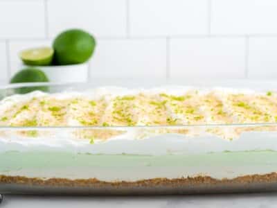 Side view of key lime lush in a 9x13 dish showing the graham cracker crust, cream cheese layer, key lime filling, and whipped topping garnished with lime zest and graham cracker crumbs