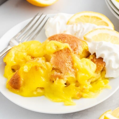 Close-up of Lemon Cobbler with glossy lemon filling and soft golden cobbler topping on a white plate, served with whipped cream and lemon slices.