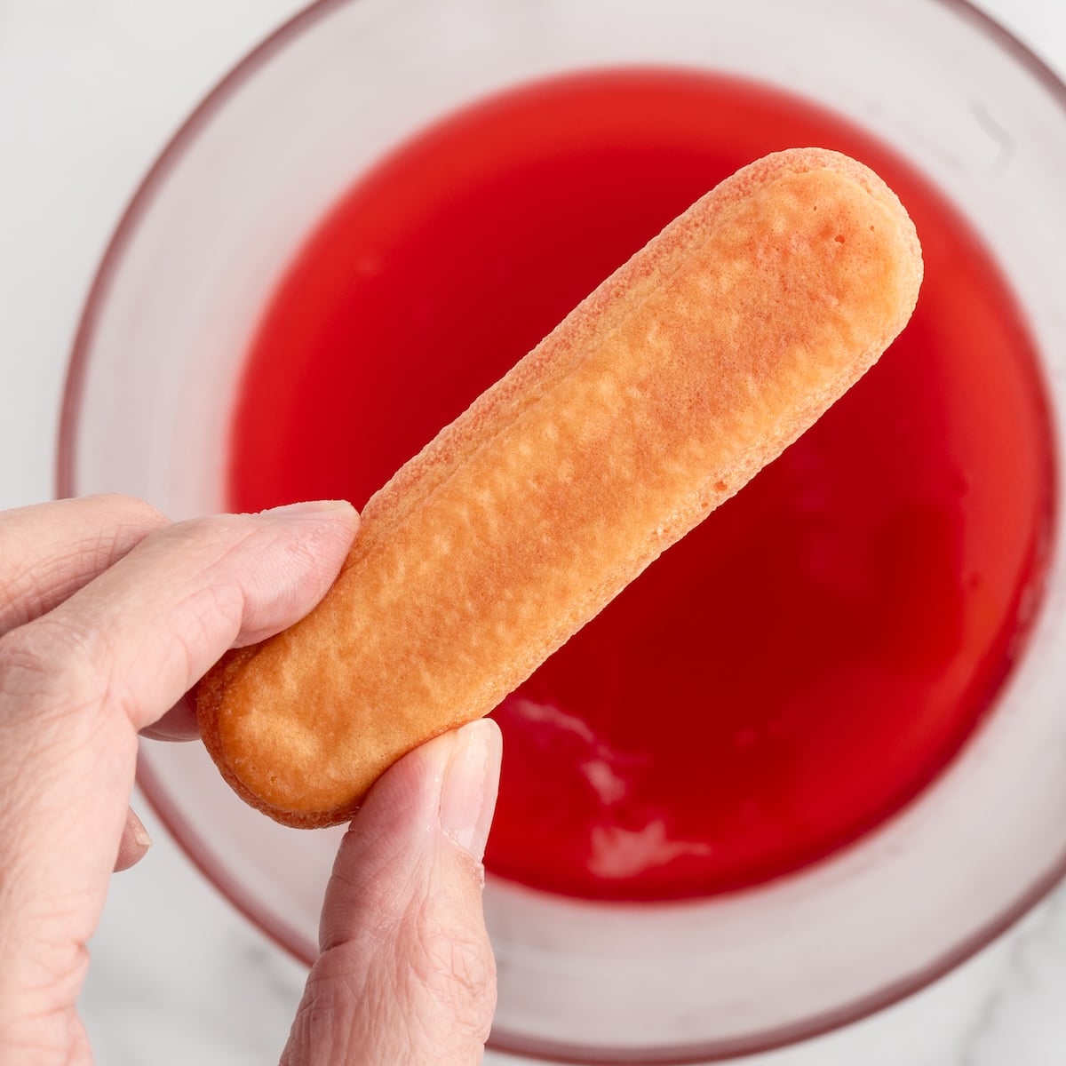 Hand holding a ladyfinger cookie above a bowl of bright raspberry syrup, preparing to dip it before layering the raspberry tiramisu