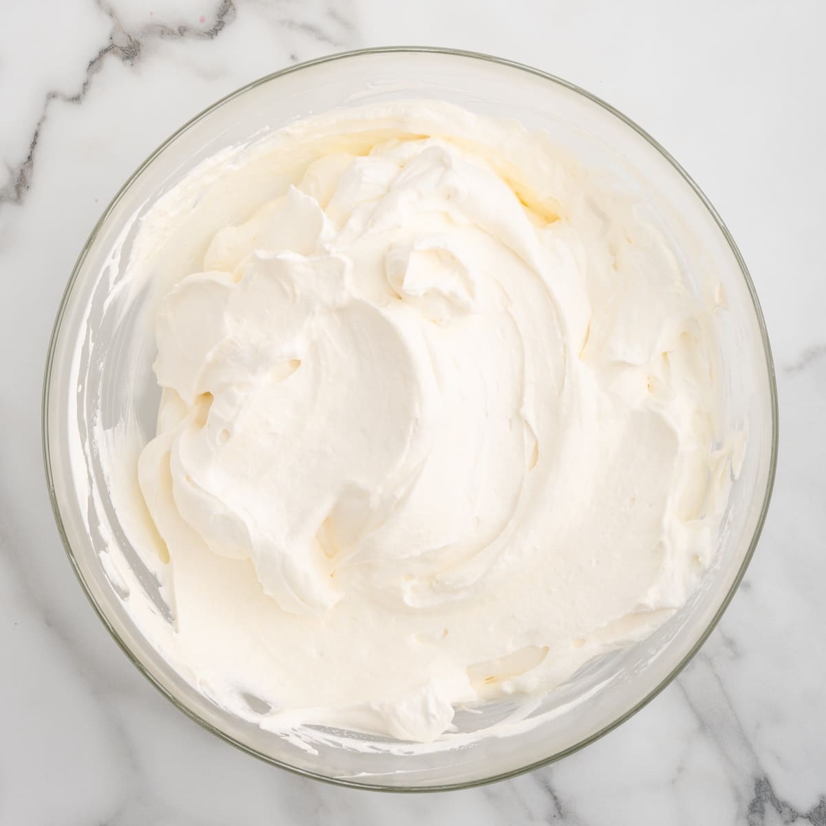 A clear glass bowl on a grey marble countertop contains combined mascarpone filling to make raspberry tiramisu