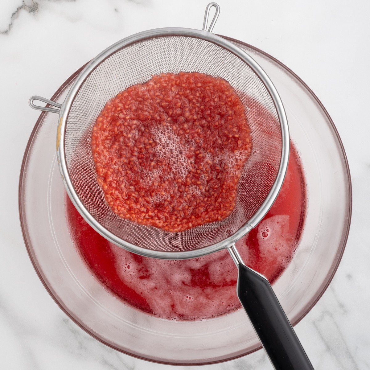 Fine mesh strainer holding mashed raspberries over a glass bowl, with bright red raspberry puree being strained to remove seeds.
