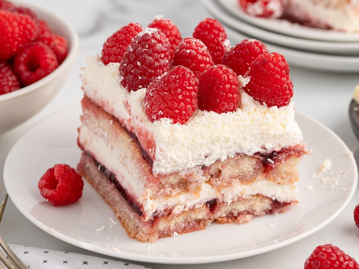 Slice of Raspberry Tiramisu on a white plate with layers of mascarpone cream, raspberry preserves, ladyfingers, grated white chocolate, and fresh raspberries.