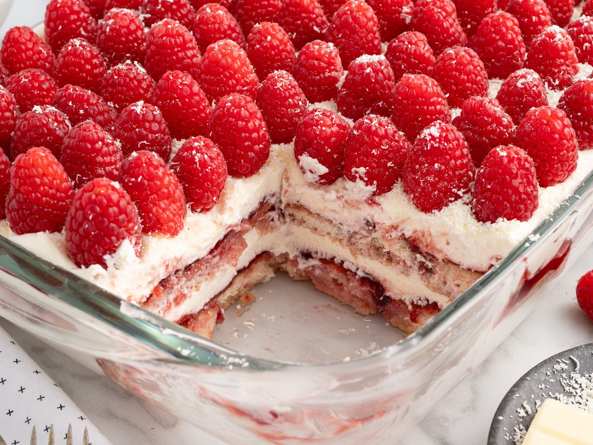 Overhead view of Raspberry Tiramisu in a glass baking dish topped with fresh raspberries and white chocolate shavings, revealing layers of mascarpone cream, raspberry preserves, and raspberry-soaked ladyfingers.
