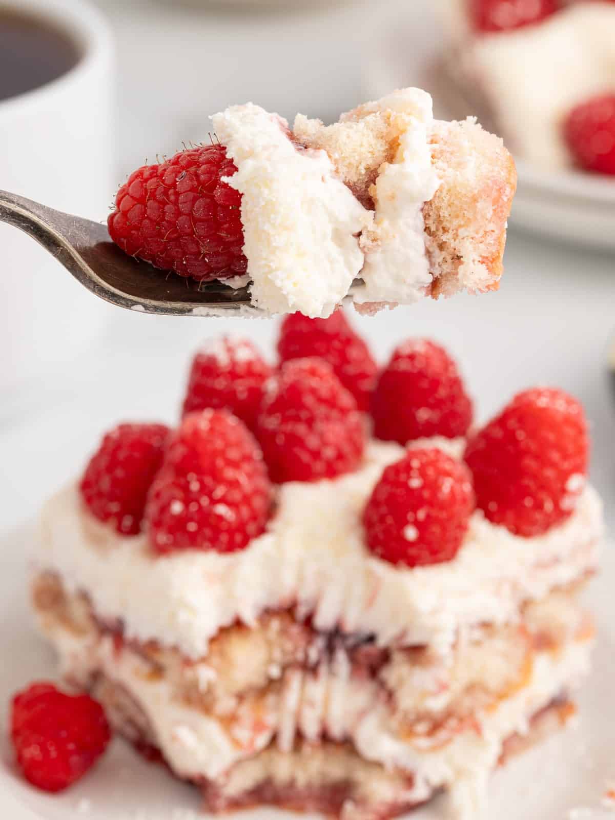 Close-up of a spoonful of Raspberry Tiramisu with fresh raspberry, fluffy mascarpone filling, raspberry preserves, and soft ladyfinger layers, with the finished dessert blurred in the background.