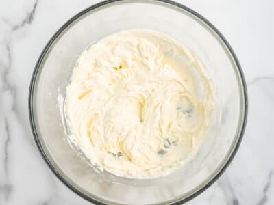 Whipped cream cheese mixture in a clear glass mixing bowl, prepared as the base for shrimp dip. The creamy mixture is smooth and fluffy, sitting on a white marble countertop under bright lighting.