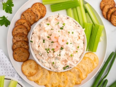 Creamy shrimp dip served in a white bowl on a platter with crackers and fresh celery sticks. The chilled seafood dip is garnished with small shrimp and chopped herbs, making it a party-ready appetizer spread.
