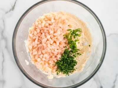 Cream cheese shrimp dip in a glass mixing bowl with small pink shrimp, chopped green onions, and fresh herbs added on top before mixing. The bowl sits on a white marble surface in bright natural light.