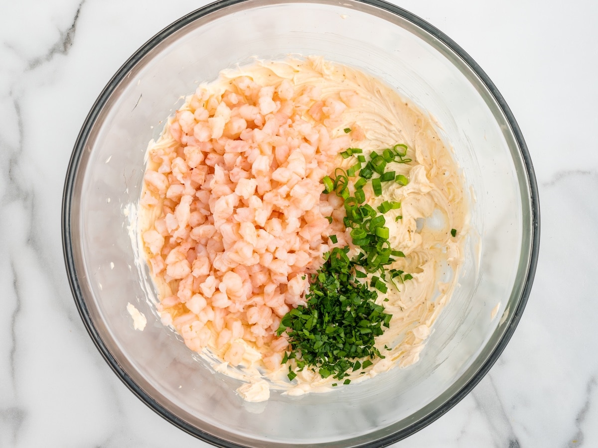 Cream cheese shrimp dip in a glass mixing bowl with small pink shrimp, chopped green onions, and fresh herbs added on top before mixing. The bowl sits on a white marble surface in bright natural light.