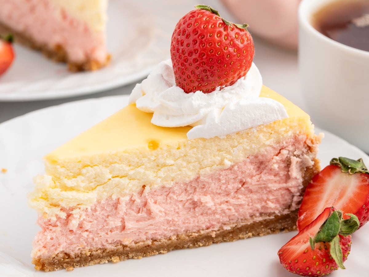 Slice of Strawberries and Cream Cheesecake showing pink strawberry cheesecake, vanilla cheesecake, graham cracker crust, whipped cream, and fresh strawberry garnish.