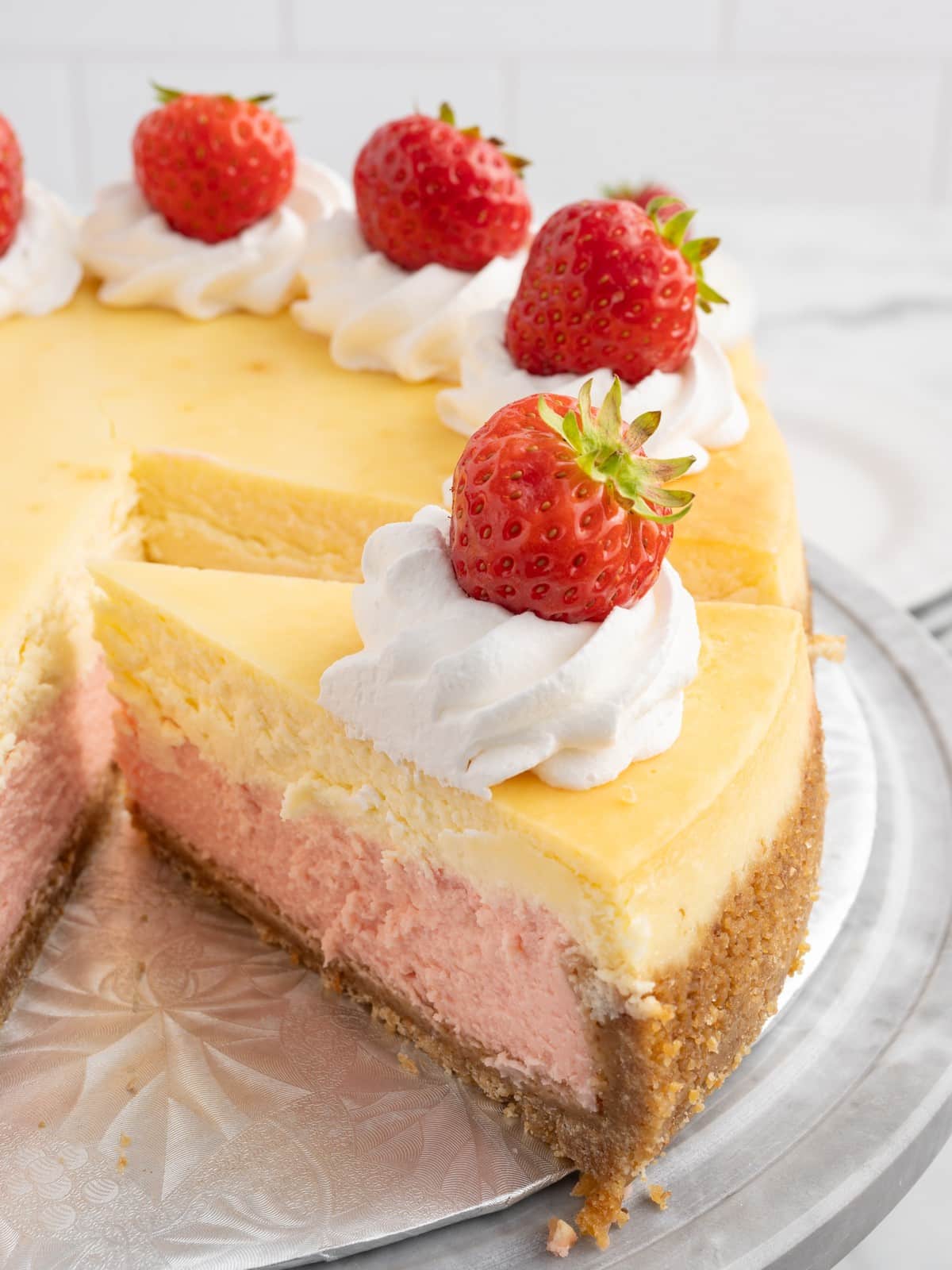 Close-up of Strawberries and Cream Cheesecake showing a thick slice removed from the cake, with a graham cracker crust, pink strawberry layer, vanilla cheesecake, whipped cream, and fresh strawberries.