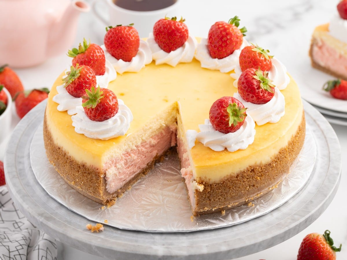 Close-up of Strawberries and Cream Cheesecake topped with whipped cream and fresh strawberries, highlighting the creamy pink strawberry cheesecake layer and buttery graham cracker crust.