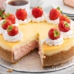 Strawberries and Cream Cheesecake topped with whipped cream and fresh strawberries, with one slice removed to reveal the creamy pink strawberry cheesecake layer and graham cracker crust.