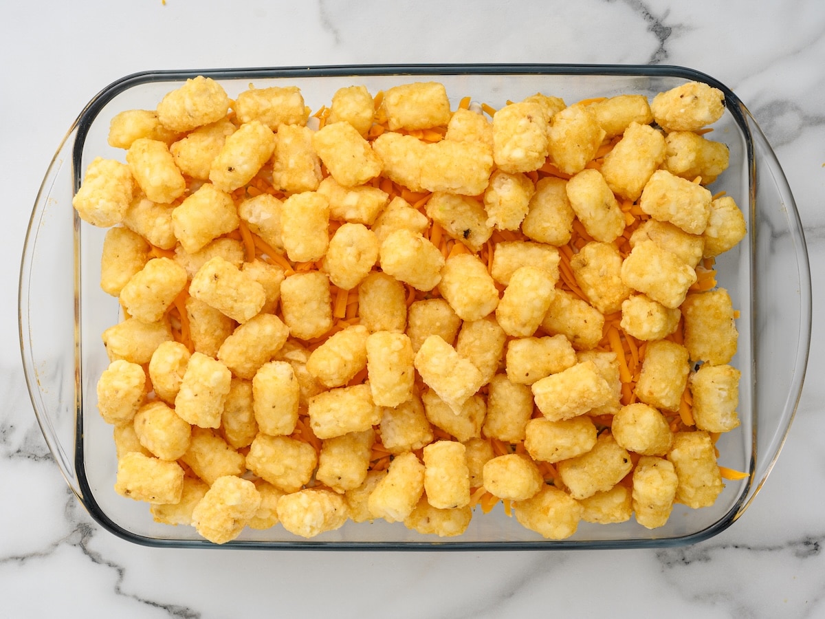 Frozen tater tots arranged in a single layer over shredded cheddar, ready to bake.