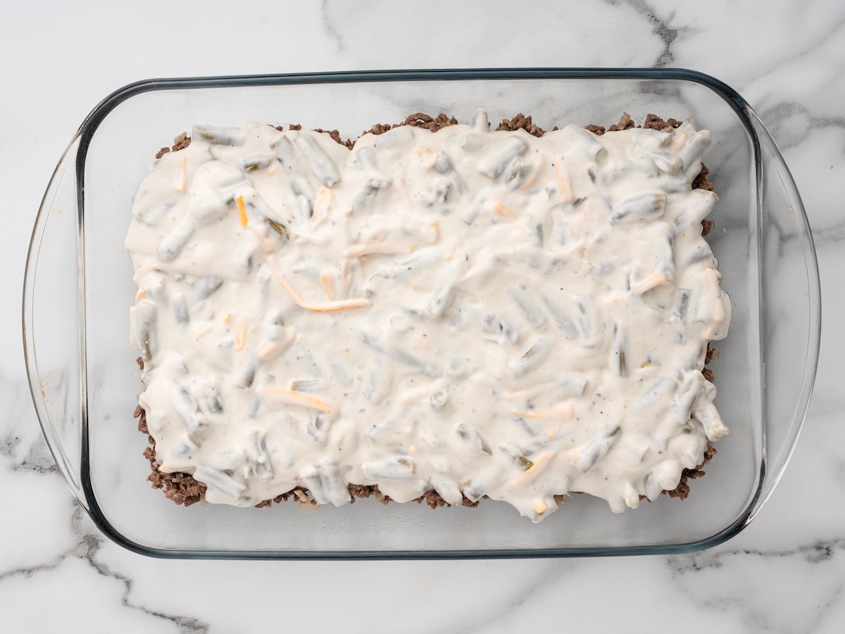 Creamy sauce with green beans and shredded cheese spooned over the ground beef layer in the baking dish.