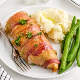 Bacon Wrapped Chicken served with creamy mashed potatoes and fresh green beans, featuring juicy chicken breasts wrapped in crispy bacon and garnished with chopped parsley on a white dinner plate.