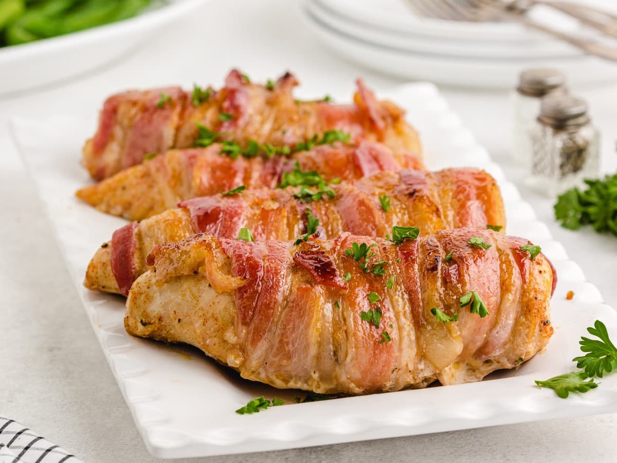 Platter of oven-baked Bacon Wrapped Chicken with crispy bacon wrapped around seasoned chicken breasts, garnished with fresh parsley and ready to serve for a family dinner.