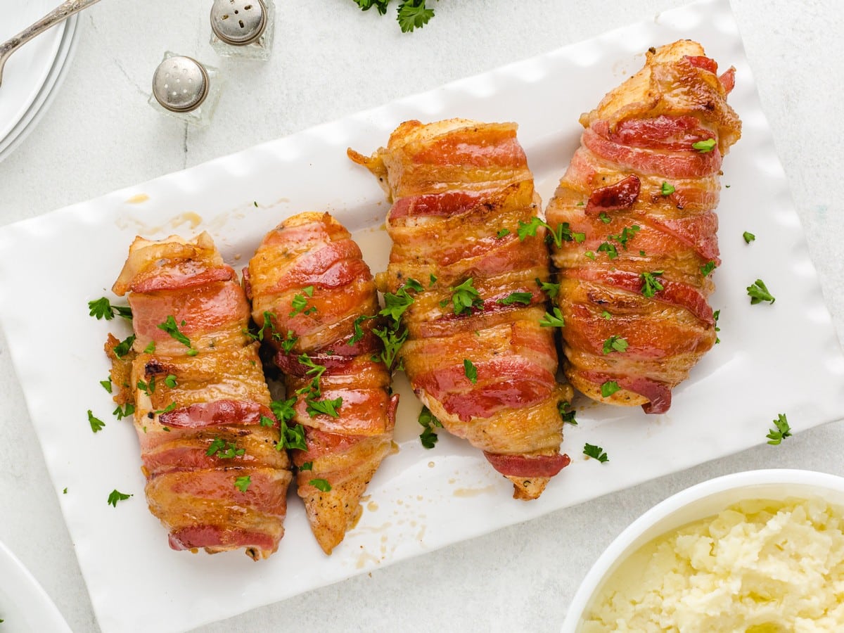 Overhead view of Bacon Wrapped Chicken arranged on a white serving platter, featuring juicy chicken breasts wrapped in crispy bacon, brushed with a sweet maple glaze, and finished with fresh parsley.