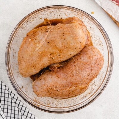 Raw boneless skinless chicken breasts coated in a maple syrup, brown sugar, and spice marinade in a glass mixing bowl before being wrapped in bacon for Bacon Wrapped Chicken.