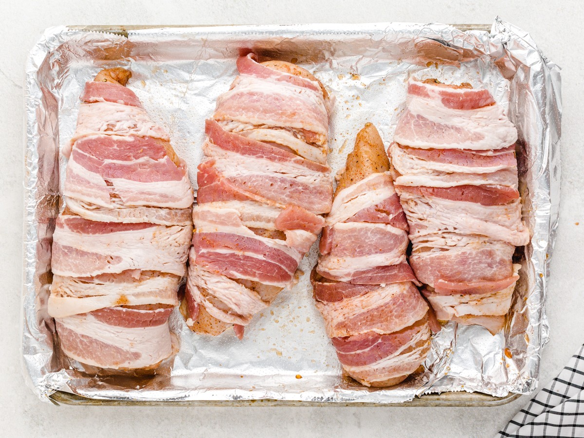 Overhead view of seasoned chicken breasts wrapped tightly with strips of bacon and arranged on a foil-lined baking sheet, ready to bake for Bacon Wrapped Chicken.