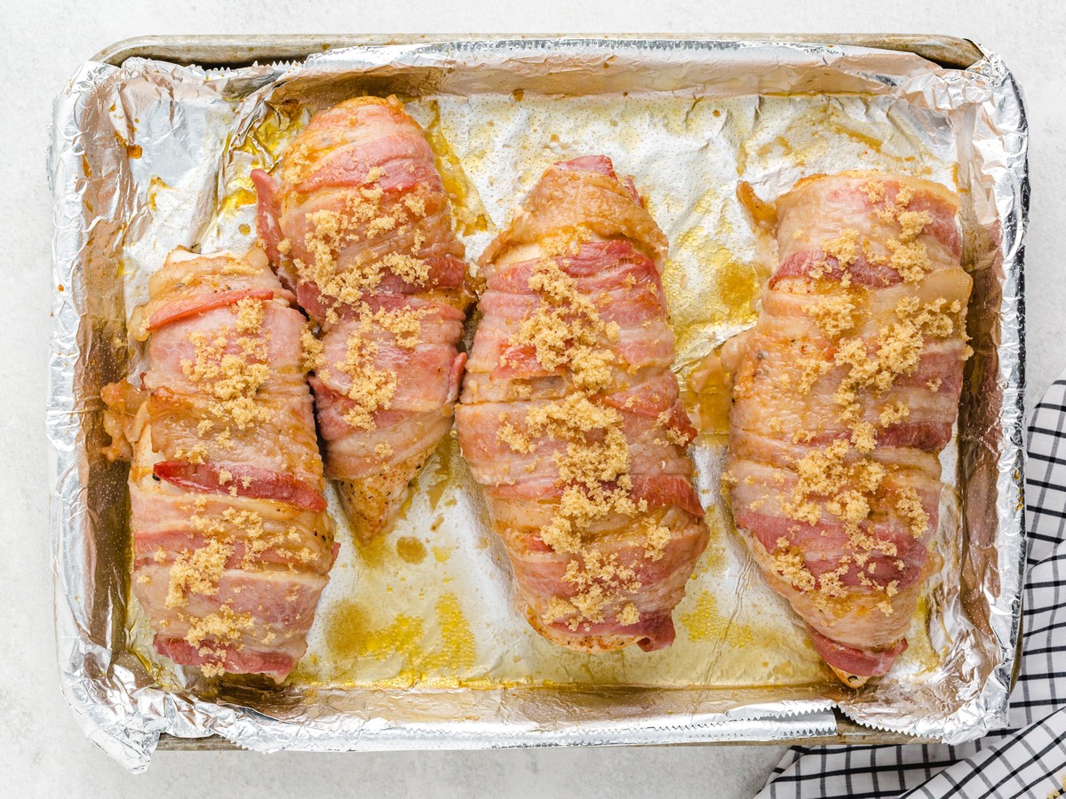 Overhead view of bacon-wrapped chicken breasts on a foil-lined baking sheet sprinkled with brown sugar before baking for sweet and savory Bacon Wrapped Chicken.