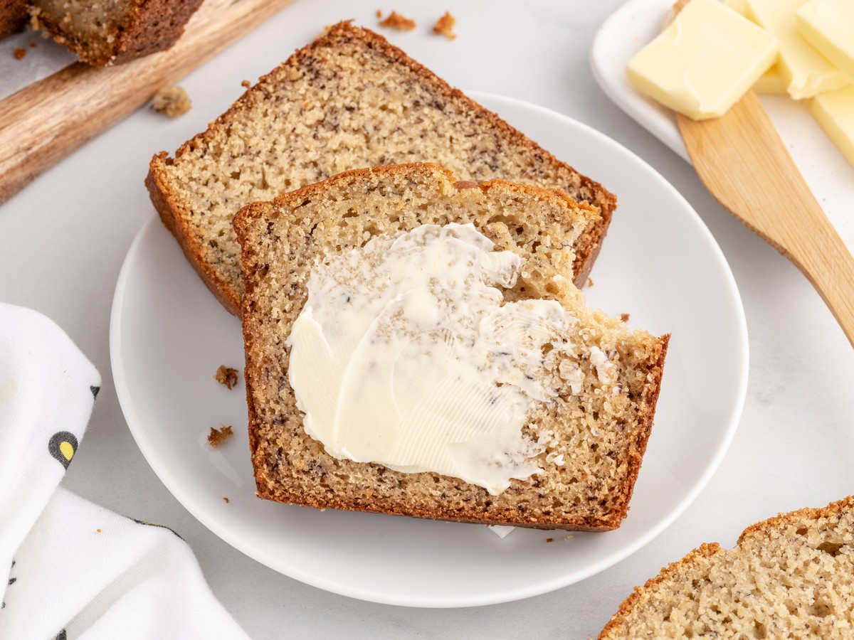 Slice of moist homemade Banana Bread generously spread with butter on a white plate, with additional slices and butter cubes in the background for a cozy breakfast serving.