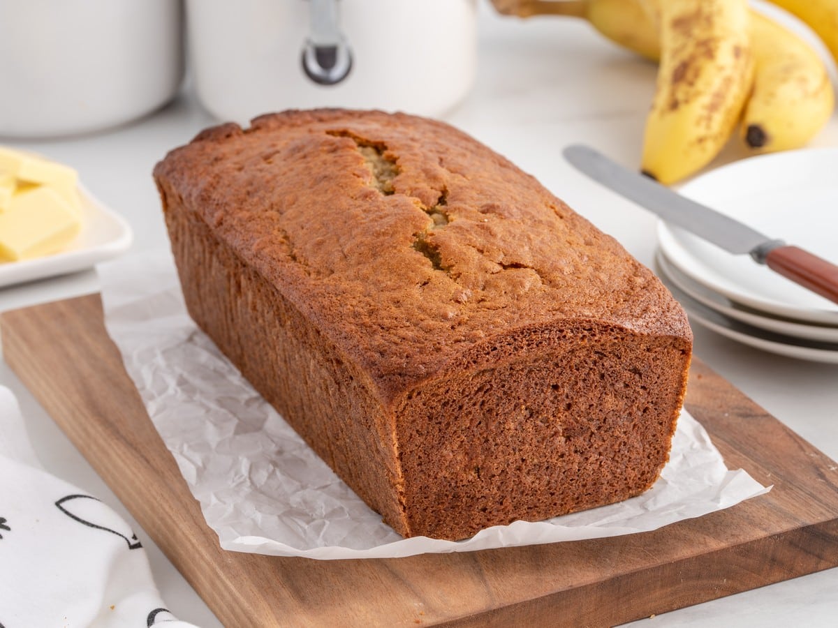 Whole homemade Banana Bread loaf with a golden brown crust cooling on parchment paper over a wooden cutting board, ready to slice and serve.