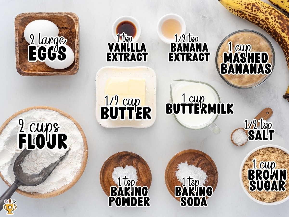 Ingredients for homemade Banana Bread arranged on a white background with text labels showing 2 large eggs, 2 cups flour, 1 cup mashed bananas, 1 cup buttermilk, 1 cup brown sugar, ½ cup butter, 1 tsp vanilla extract, ½ tsp banana extract, 1 tsp baking powder, 1 tsp baking soda, and ½ tsp salt.