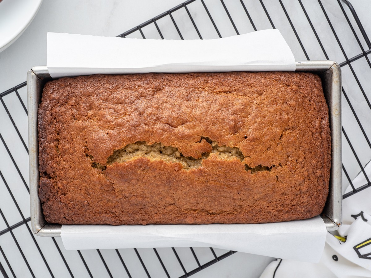 Freshly baked Banana Bread cooling in a loaf pan on a wire rack, showing a golden brown crust with the signature crack across the top.