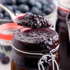 Close-up of a spoonful of glossy Blueberry Butter over a filled jar with fresh blueberries behind it.