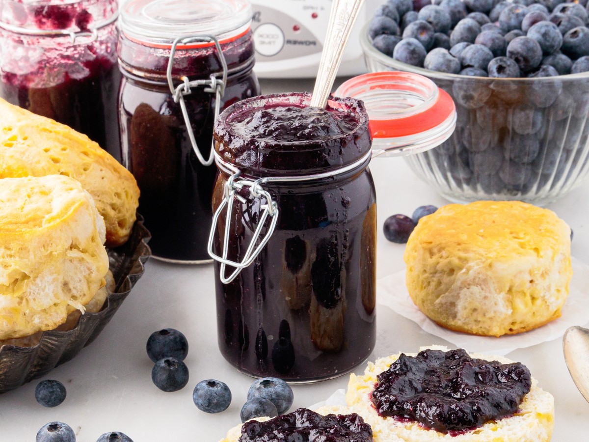 Jars of homemade Blueberry Butter served with fresh blueberries and biscuits, with dark blueberry spread on split biscuits.