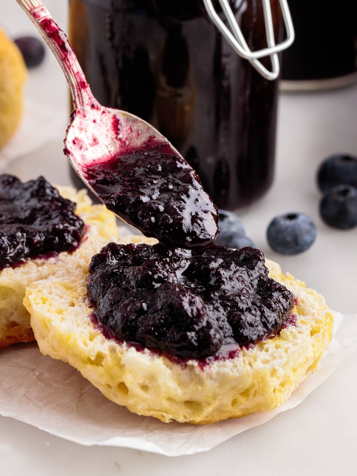 Spoon spreading thick homemade Blueberry Butter onto a warm split biscuit with fresh blueberries and a jar of blueberry butter in the background.