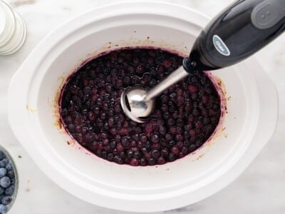 Cooked blueberries in the slow cooker being blended with an immersion blender to create a smooth, rich Blueberry Butter with a thick fruit spread consistency.