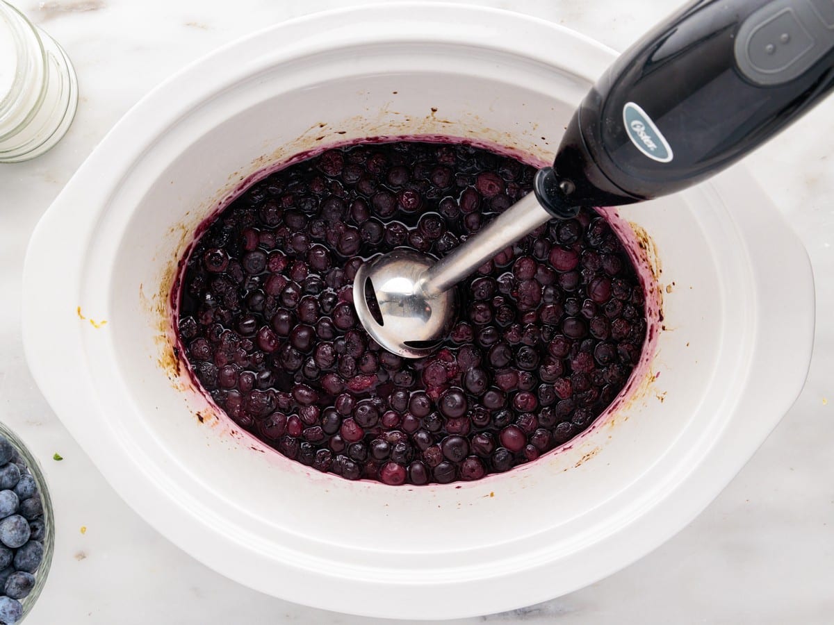 Cooked blueberries in the slow cooker being blended with an immersion blender to create a smooth, rich Blueberry Butter with a thick fruit spread consistency.