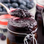 Spoon lifting a thick, glossy scoop of homemade Blueberry Butter above a clip-top glass jar, showcasing the rich texture of the slow cooker fruit spread with fresh blueberries and filled jars in the background.